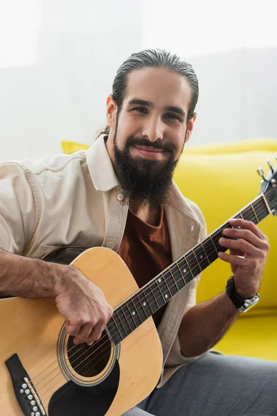 Позитивний іспаномовний чоловік дивиться на камеру, граючи на гітарі вдома — стокове фото