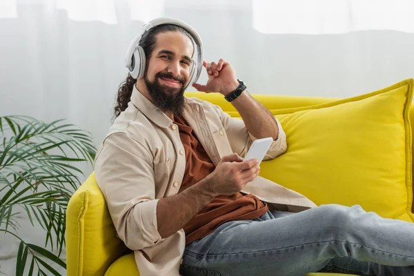 Cheerful hispanic man with smartphone looking at camera while listening music in headphones on yellow couch — Stock Photo