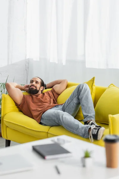 Vista completa del hombre latino descansando en el sofá amarillo cerca de café borroso para ir y portátil - foto de stock