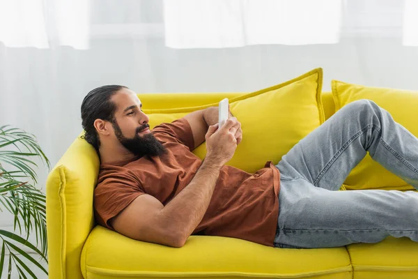 Uomo ispanico chatta su smartphone mentre riposa su un divano giallo a casa — Foto stock