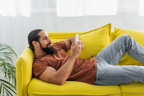 Spanischer Mann plaudert auf Smartphone, während er sich zu Hause auf gelbem Sofa ausruht — Stockfoto