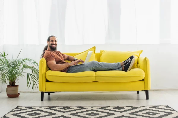 Cheerful hispanic man smiling at camera while using laptop on yellow sofa — Stock Photo
