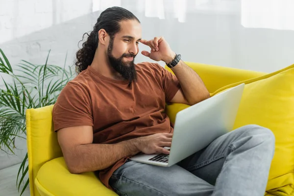 Positive and thoughtful hispanic man looking at laptop while sitting on sofa at home — Stock Photo