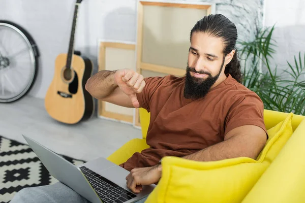 Hispanic man showing dislike gesture while using laptop on couch near blurred guitar and canvases — Stock Photo