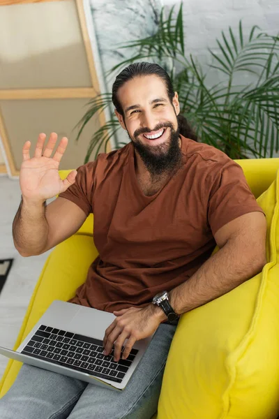 Felice uomo latino agitando la mano davanti alla macchina fotografica mentre seduto sul divano con il computer portatile — Foto stock