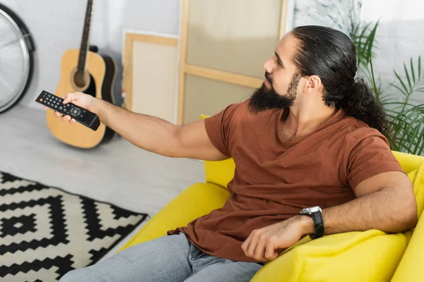 Vista de ángulo alto del hombre hispano haciendo clic en los canales de televisión cerca borrosa guitarra acústica y lienzos - foto de stock