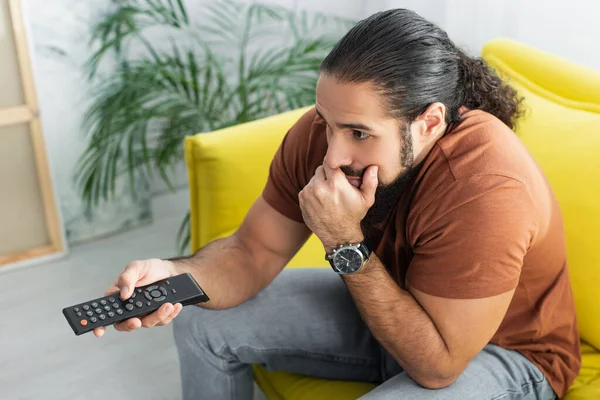 Hochwinkel-Ansicht eines nachdenklichen hispanischen Mannes mit Fernbedienung, der zu Hause Filme anschaut — Stockfoto