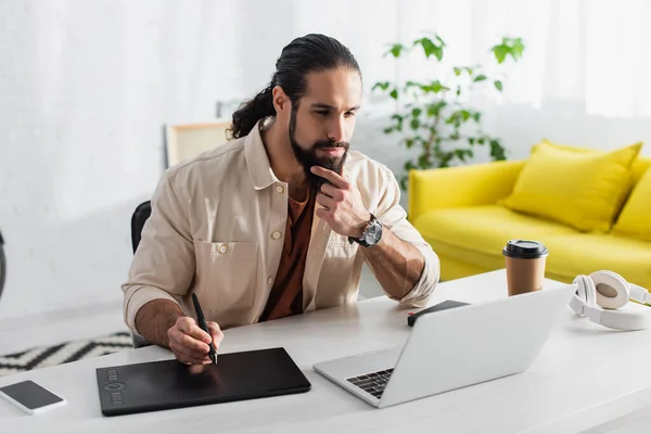 Retouche hispanique réfléchie toucher la barbe tout en travaillant avec ordinateur portable et tablette graphique à la maison — Photo de stock