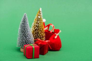 Süslü gümüş ve altın noel ağaçları hediye kutularının yanında, çuval ve alışveriş çantası ve yeşil şapkalı Noel Baba