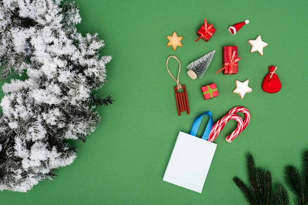 top view of shopping bag with candy canes near christmas baubles and pine wreath with decorative snow on green background