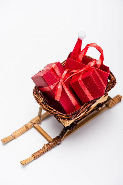 red gift boxes and shopping bag with santa hat in wicker basket on decorative sled on white background