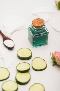 Taze tonik şişesi, salatalık dilimleri, çay gülleri, kozmetik kremalı kaşık ve beyaz bulanık arka planda pamuk pedleri.