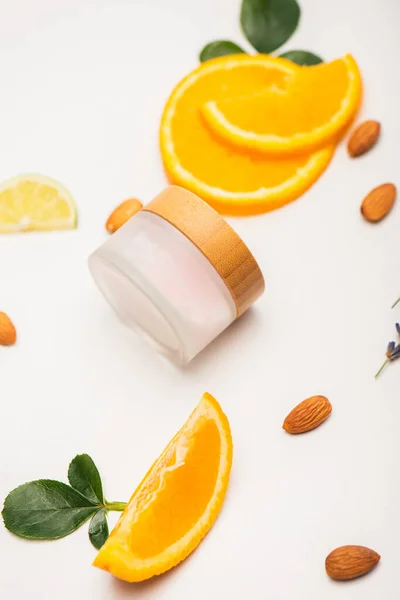 container of homemade cosmetic cream near fresh orange slices, almonds and rose leaves on white