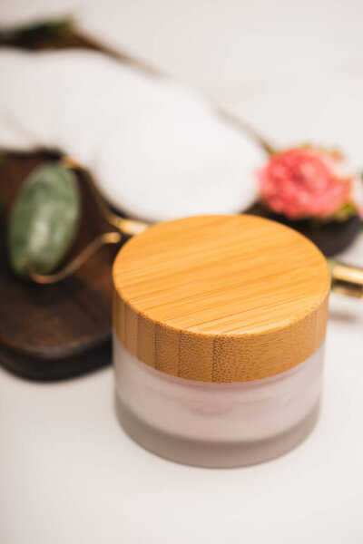 container with cosmetic cream near jade roller and cotton pads on chopping board on white blurred background