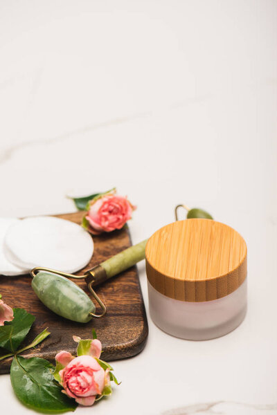 tea roses and jade roller on chopping board near homemade cosmetic cream on white blurred background