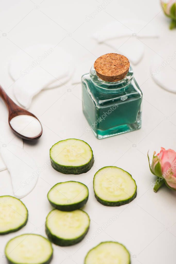 corked bottle of refreshing tonic, cucumber slices, tea roses, spoon with cosmetic cream and cotton pads on white blurred background