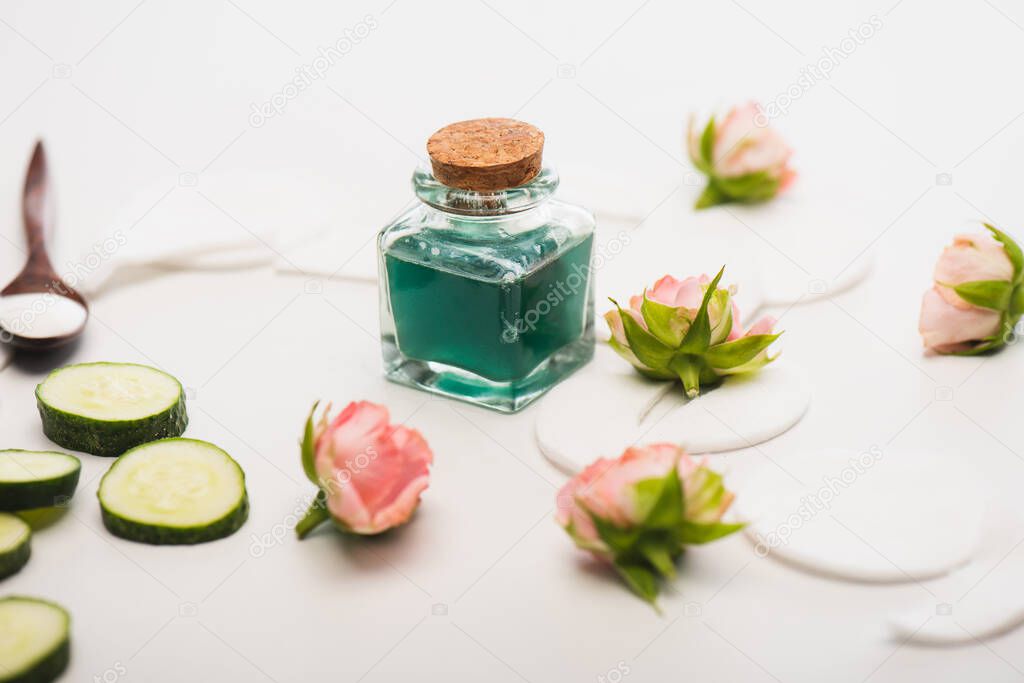 corked bottle of homemade tonic near tea roses, spoon with cosmetic cream on white blurred background