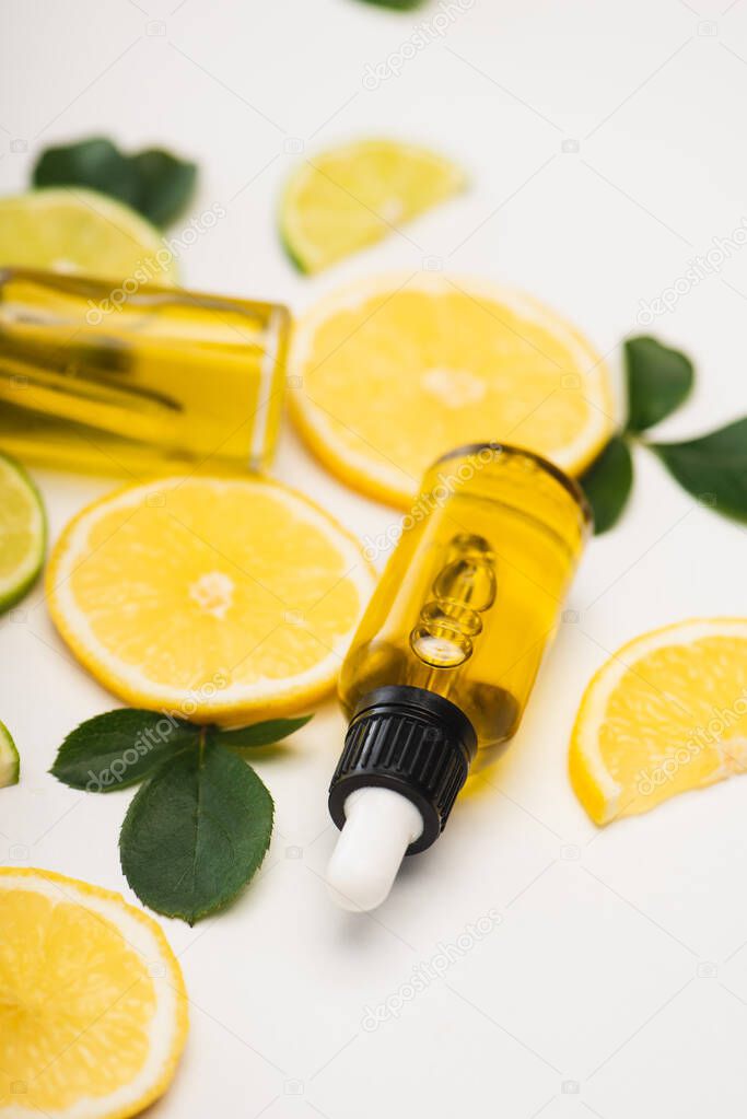 selective focus of lemon and lime slices near bottles of citrus essential oil and rose leaves on blurred background
