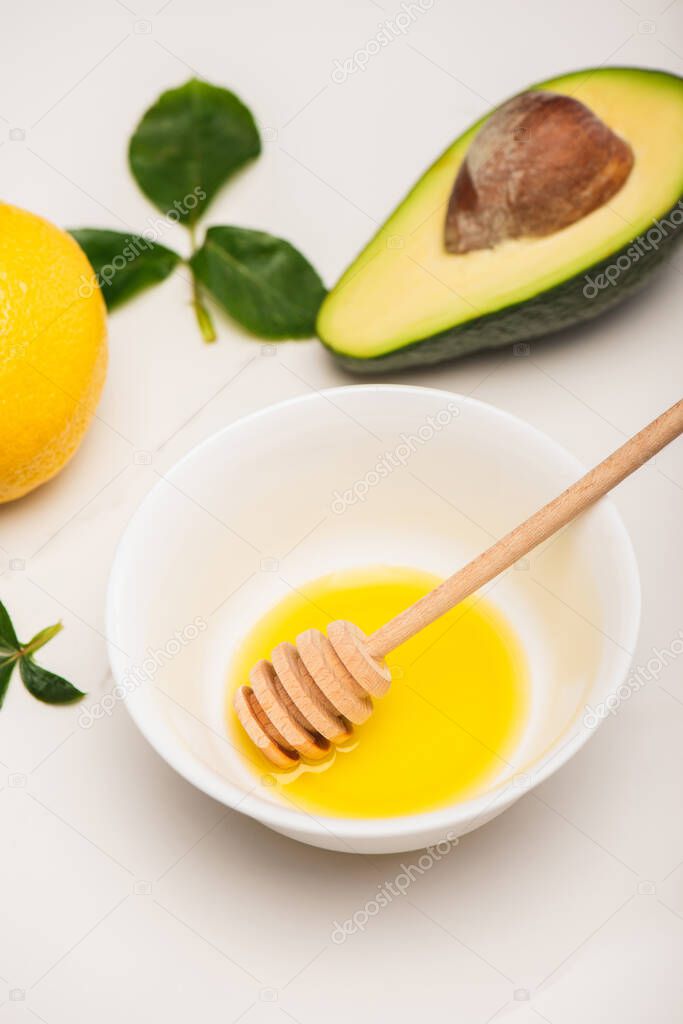 bowl with honey and dipper, fresh avocado, lemon and rose leaves on white