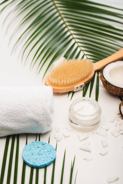 coconut half, cosmetic cream, towel, massage brush and sponge near palm leaves on white