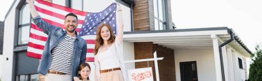Mutlu çift ve kızı kameraya bakarken Amerikan bayrağı ellerinde satılan harfler, ev ve afiş ile