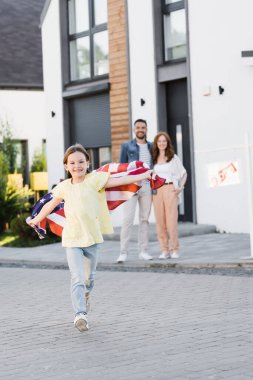 Arka planda bulanık ebeveynler sarılıp kameraya bakarken Amerikan bayrağıyla ileri doğru koşan mutlu bir kız.