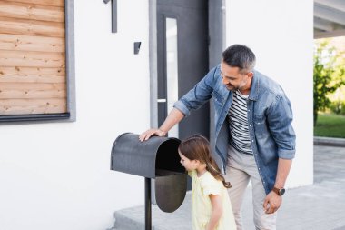 Smiling father standing near daughter looking in empty mailbox near house clipart
