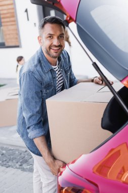 Happy man looking at camera while taking carton box from car trunk on blurred background clipart