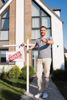 Tam uzunlukta gülümseyen bir adam, arka planda modern evle birlikte satılan harflerle tabelanın yanında dururken anahtar gösteriyor.