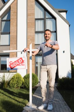 Full length of smiling man showing key while standing near sign with sold lettering with modern house on background clipart