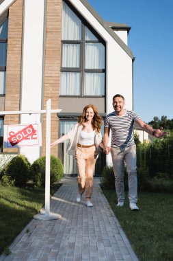 Cheerful couple holding hands while running forward near sign with sold lettering and modern house clipart