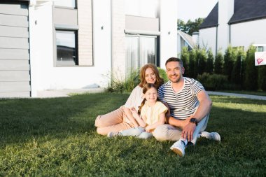 Modern evin yakınındaki çimlerde oturan mutlu aile kameraya bakıyor.