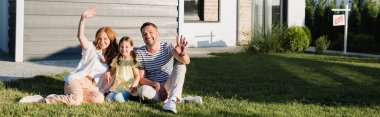 Happy parents with waving hands and daughter sitting on lawn and looking at camera near house, banner clipart