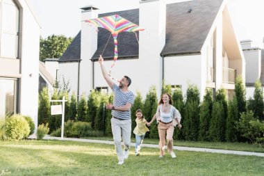 Full length of happy man flying kite near woman and girl on lawn near houses clipart
