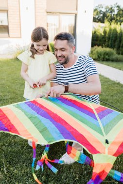Full length of smiling daughter standing near father assembling kite on lawn with blurred house on background clipart