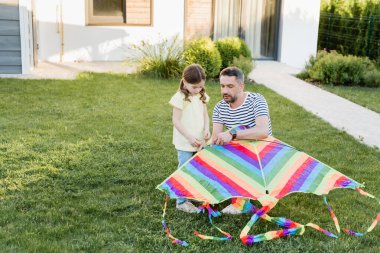 Full length of daughter standing near father assembling kite on lawn with house on background clipart