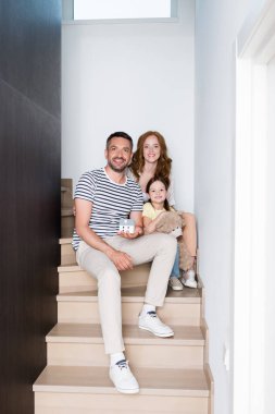 Oyuncak ayı ve ev heykelciğiyle mutlu bir aile evdeki merdivenlerde otururken kameraya bakıyor.