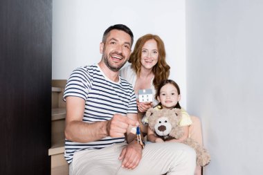 Evin heykelciği ve evinde oyuncak ayısıyla merdivenlerde oturan mutlu adam anahtarları gösteriyor.