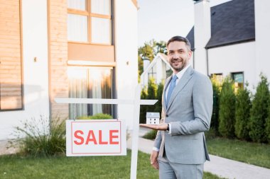 Smiling real estate agent with house statuette looking at camera near sign with sale lettering with blurred building on background clipart