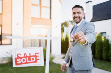 smiling broker looking at camera while showing keys near sign with sold lettering on blurred background clipart