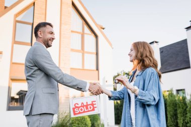 Side view of happy broker and woman with keys shaking hands near sign with sold lettering on blurred background clipart