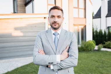 Çapraz kollu kendine güvenen simsar arka planda bulanık evleri olan kameraya bakıyor.