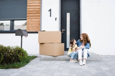 Happy mother and daughter looking at each other while sitting near carton boxes on doorstep clipart