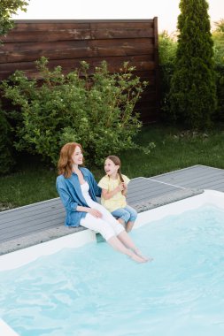 Mutlu anne ve kızın yüksek açılı manzarası arka bahçesinde çalılar ve çimlerin yanında bacaklarıyla yüzme havuzunda oturması.