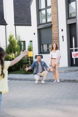 Kızıl saçlı kadın, ön planda bulanık kızına bakarken açık kollarıyla çömelmiş adamın yanında duruyor.