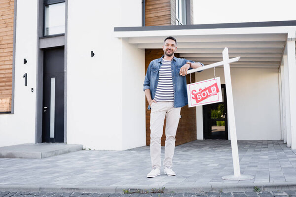 Full length of smiling man with hand in pocket leaning on sign with sold lettering near house