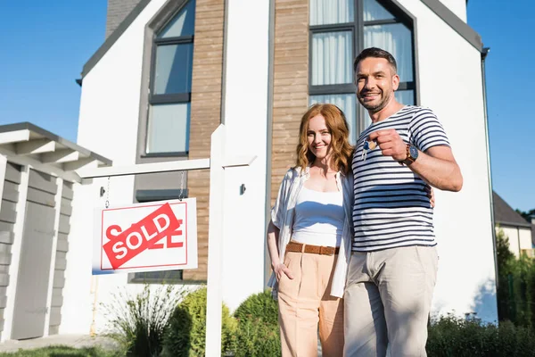 Smiling couple with keys hugging while standing near sign with sold lettering and modern house on background