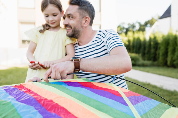 Smiling father looking at daughter while assembling kite on blurred background