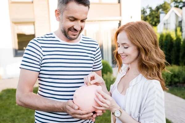 Smiling Redhead Wife Putting Coin Piggy Bank Husband Blurred Background — Stock Photo, Image
