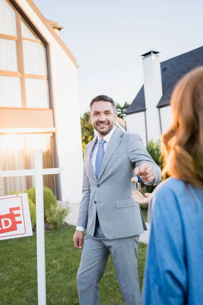 Smiling Broker Looking Camera While Giving Keys Blurred Woman Foreground — Stock Photo, Image